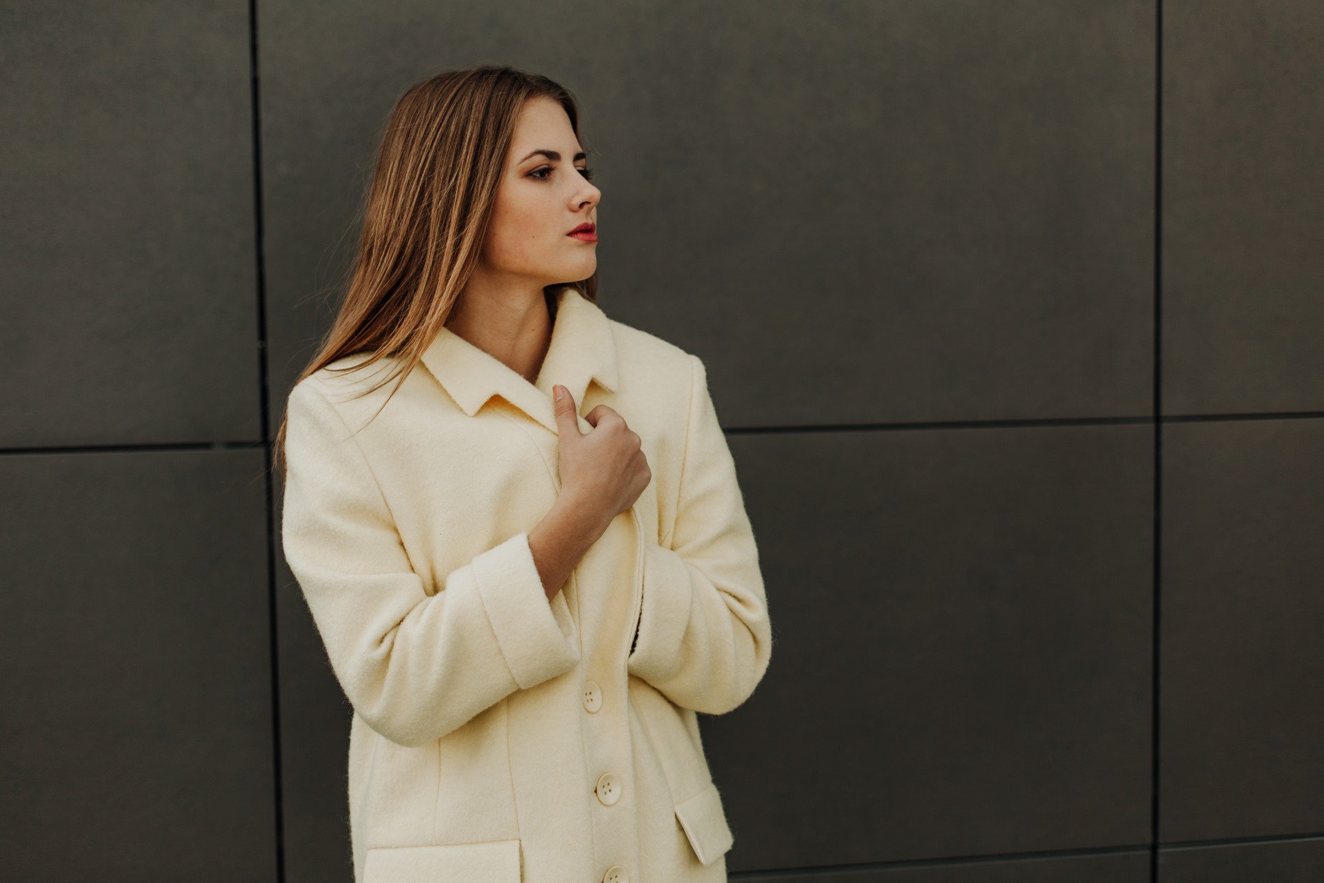 A woman in a beige coat, perfect for winter office fashion, stands against a dark gray tiled wall, looking to her right with her hands clasped near the collar.